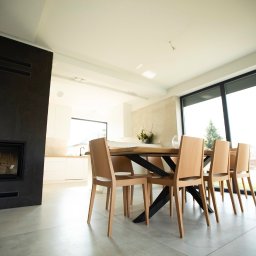 Rem-Bud Grzegorz Bernatek - Jasne wnętrze jadalni z drewnianym stołem i krzesłami, nowoczesny kominek z czarną obudową, duże okno z widokiem na zewnątrz, szara podłoga z płytek.