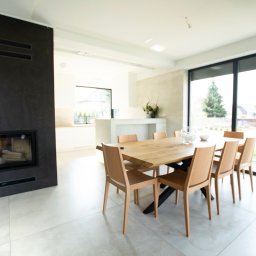 Rem-Bud Grzegorz Bernatek - Jasne wnętrze z kominkiem o ciemnej obudowie i drewnianym stołem. Minimalistyczny design, duże okno z widokiem na zieleń. Nowoczesne wykończenie.