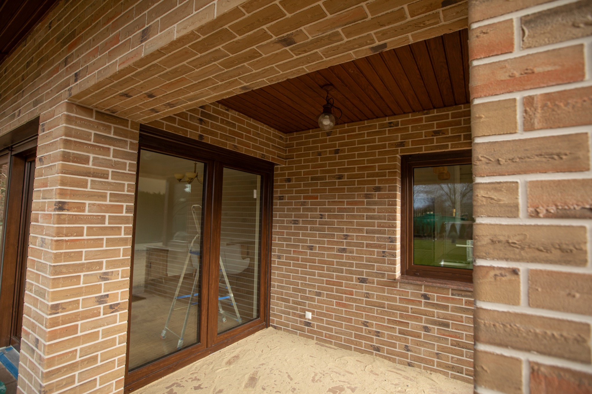 Wykończony taras z ceglaną elewacją, brązowe okna i drzwi balkonowe. Widoczna drabina wewnątrz. Podłoga pokryta piaskiem. Drewniana podbitka dachowa.