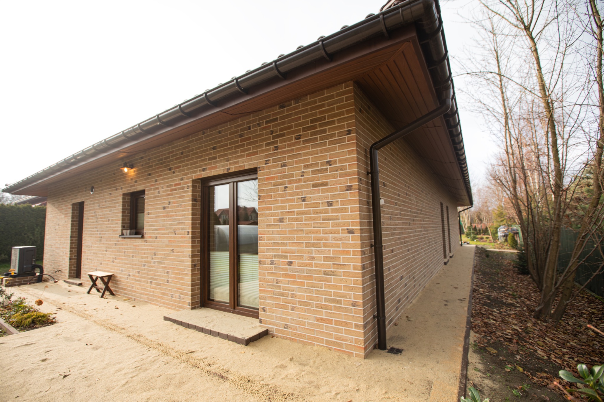Nowoczesny, parterowy dom z cegły klinkierowej w odcieniach beżu i brązu. Widoczne okna z brązowymi ramami, brązowe rynny i oświetlenie zewnętrzne. Otoczenie w stanie surowym.