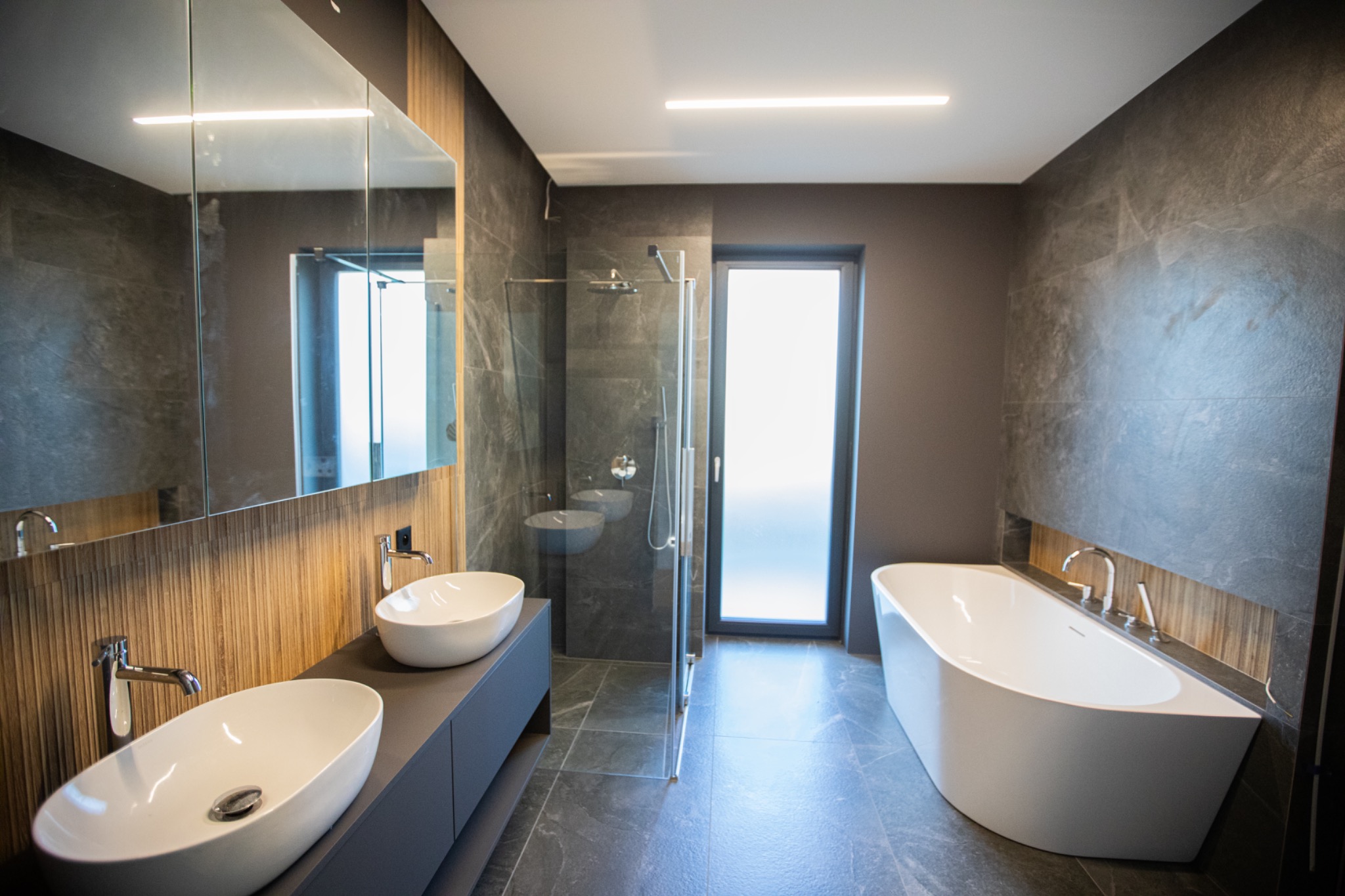 Nowoczesna łazienka z dwoma umywalkami na szafce, kabiną prysznicową ze szklaną ścianką i wolnostojącą wanną. Szare płytki imitujące kamień na ścianach i podłodze.