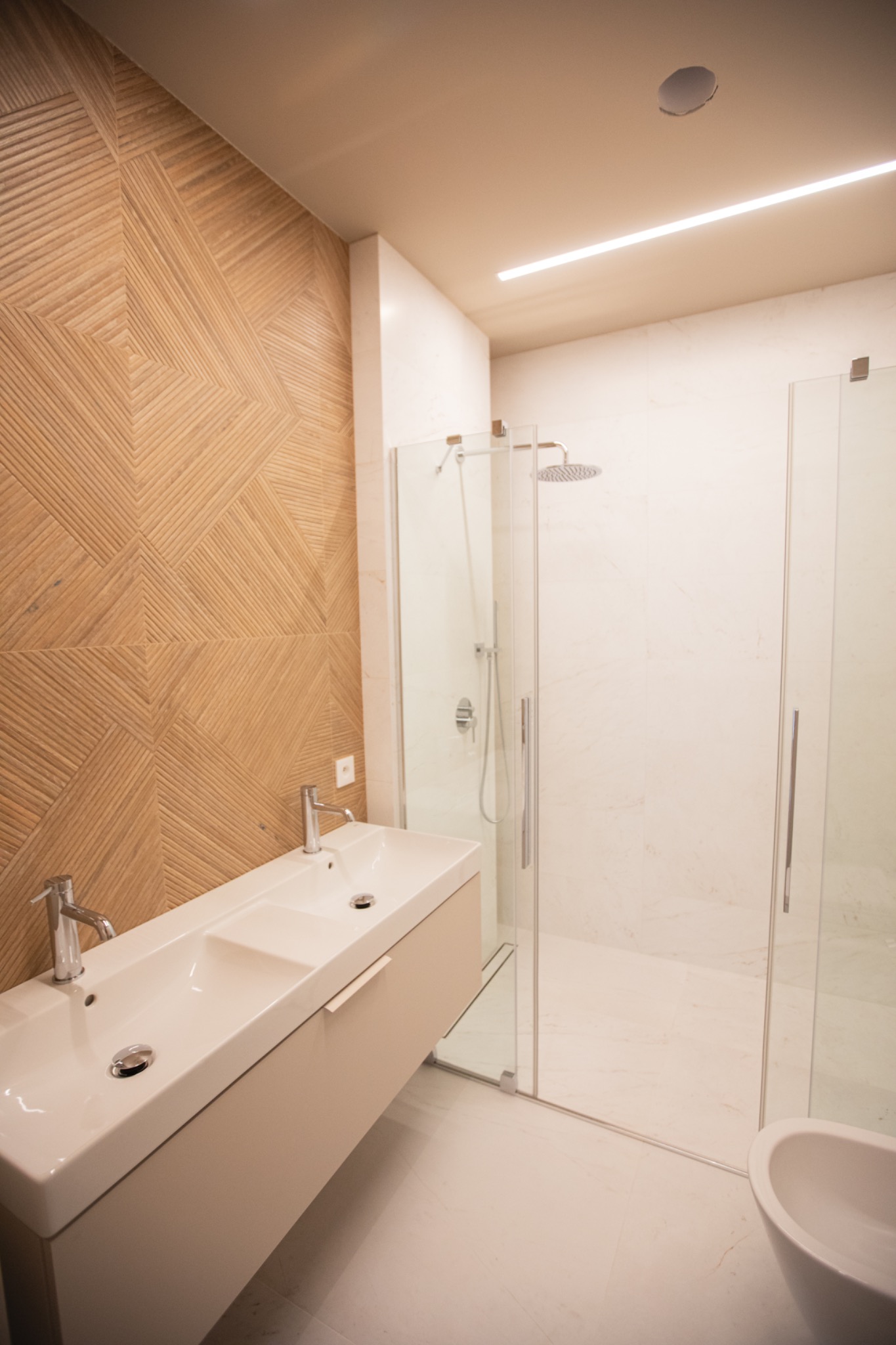 Nowoczesna łazienka z drewnianą ścianą, podwójną umywalką i kabiną prysznicową ze szklanymi drzwiami. Minimalistyczny design, jasne kolory i oświetlenie LED.