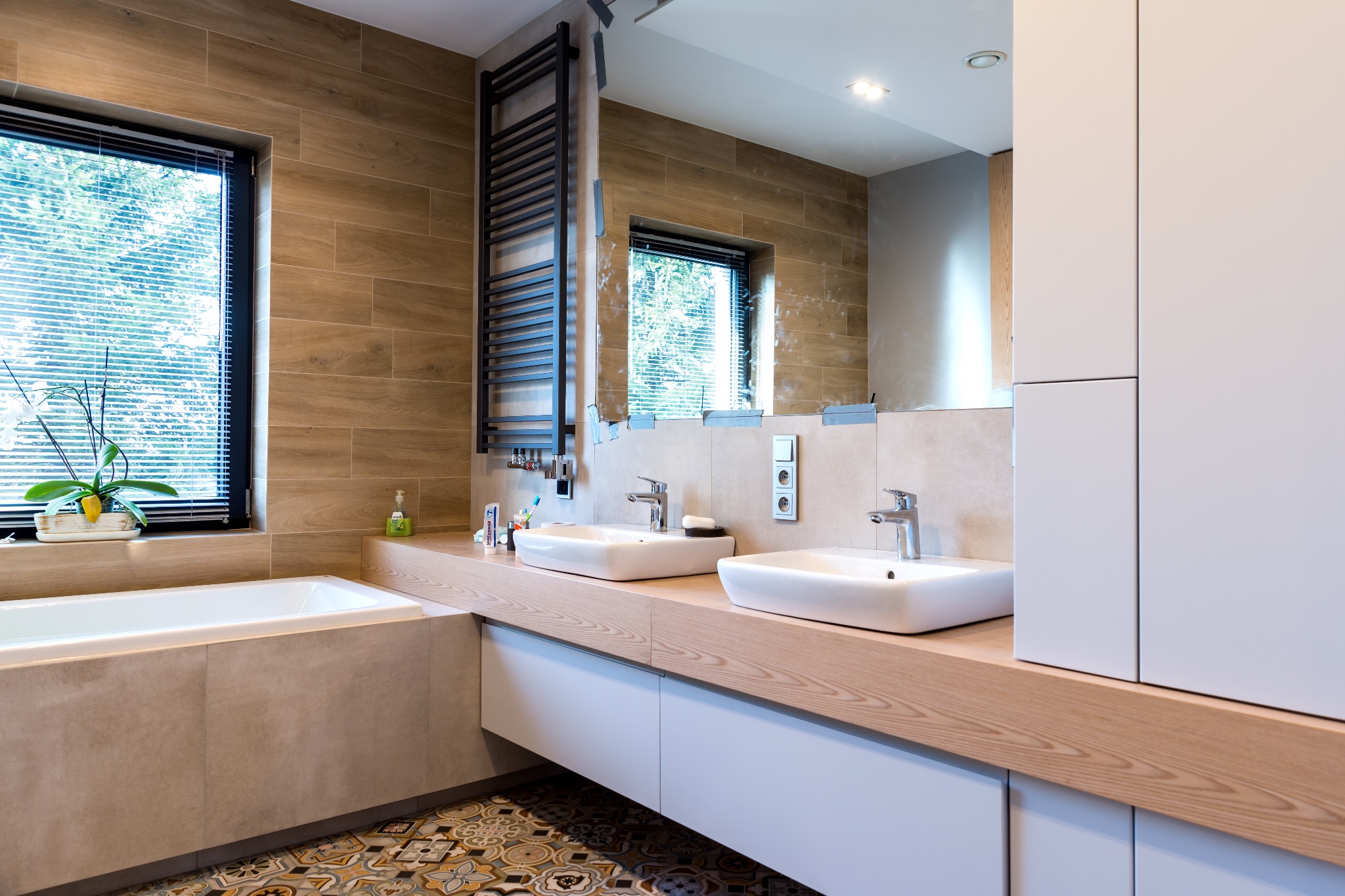 Nowoczesna łazienka z dwoma umywalkami, drewnianym blatem i wzorzystą podłogą. Na ścianach płytki imitujące drewno, grzejnik drabinkowy i wanna przy oknie.