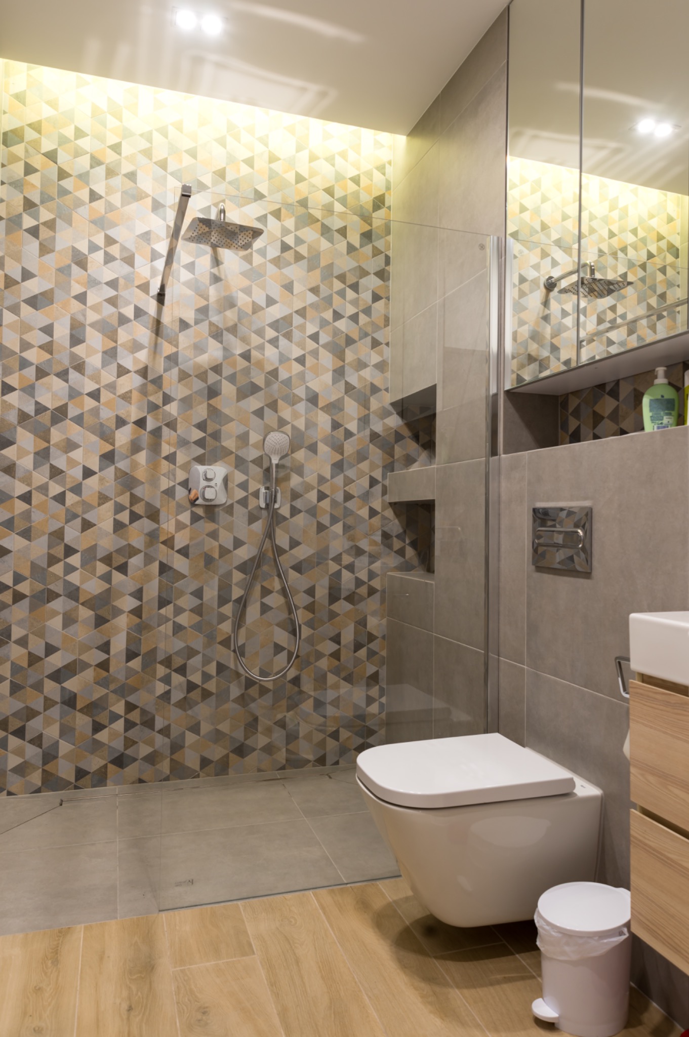 Nowoczesna łazienka z kabiną prysznicową typu walk-in, wykończona geometrycznymi płytkami w odcieniach szarości i beżu. Widoczna toaleta podwieszana i drewniana szafka.