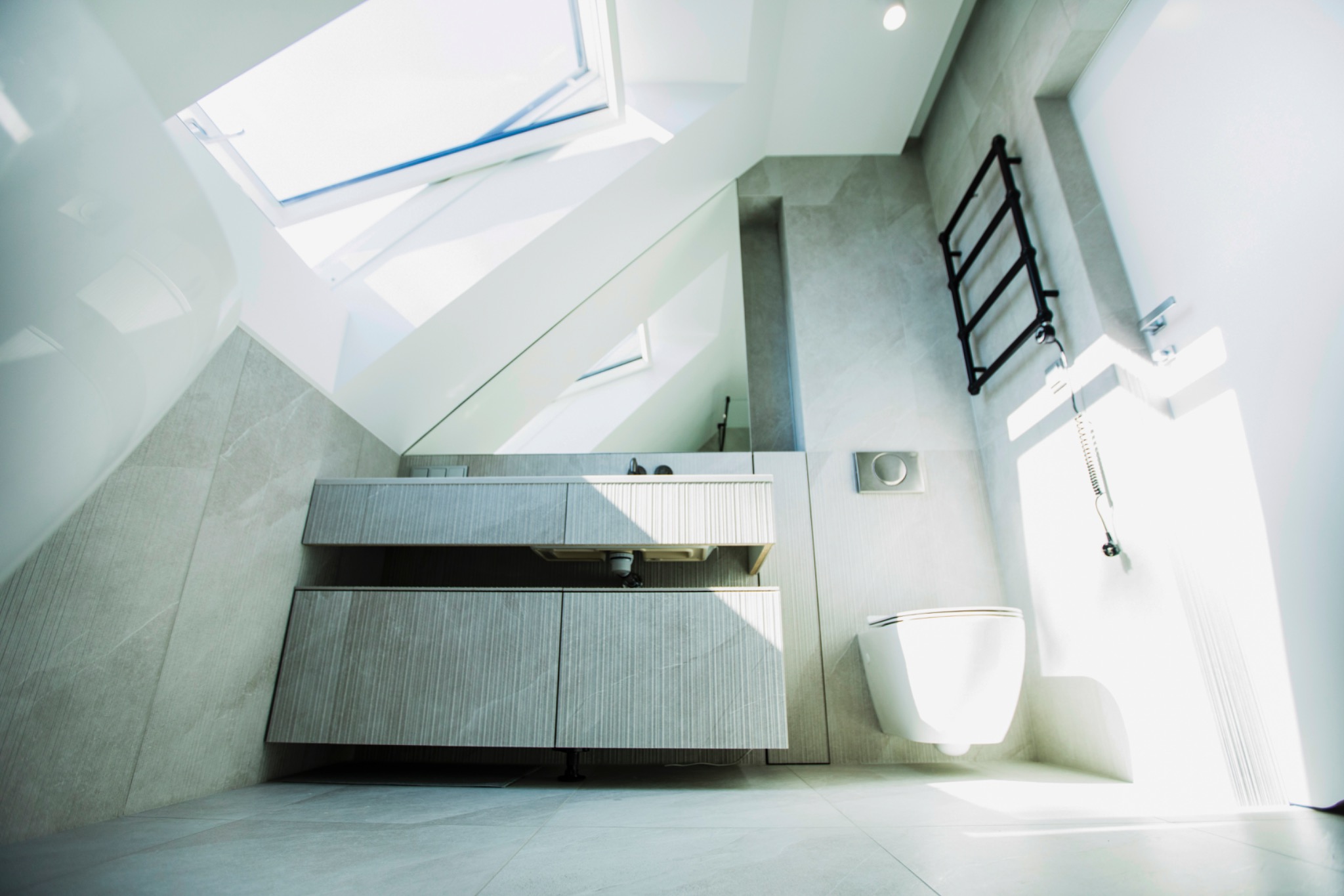 Jasna łazienka na poddaszu z oknem dachowym. Umywalka z szafką w odcieniach szarości, wisząca toaleta i grzejnik drabinkowy. Minimalistyczny design, płytki imitujące kamień.