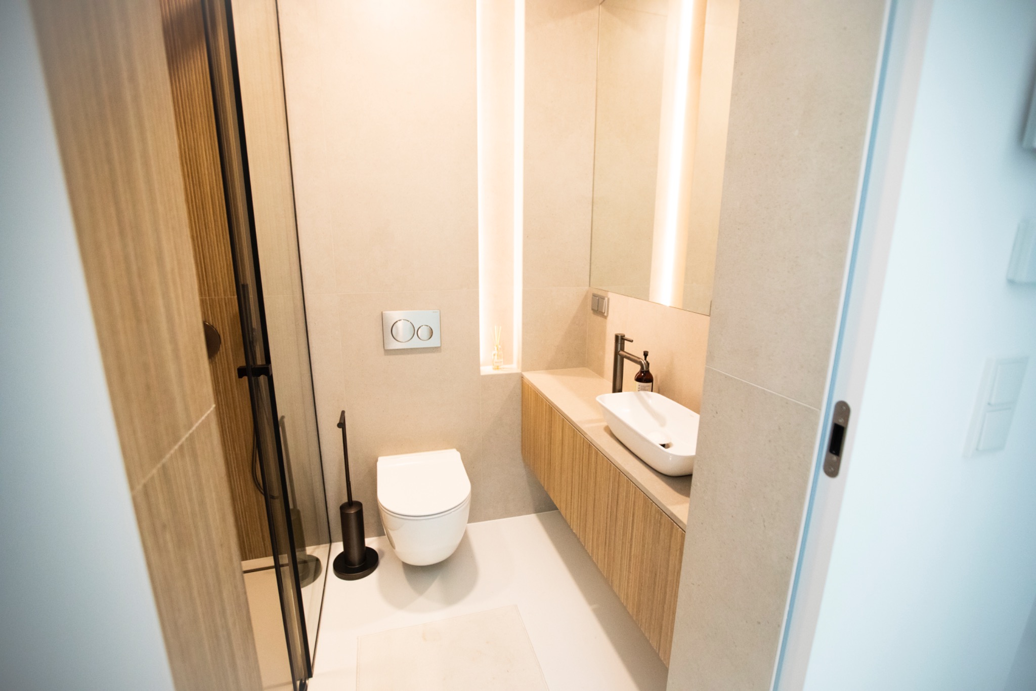 Nowoczesna łazienka w beżowej tonacji z podświetleniem LED, wiszącą toaletą i umywalką na drewnianej szafce. Widoczna kabina prysznicowa z czarnymi elementami.