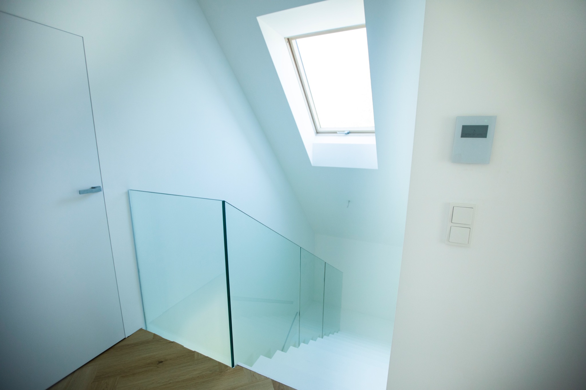 Minimalistyczne wnętrze: białe schody z szklaną balustradą, drewniana podłoga, okno dachowe i białe ściany z panelem sterowania. Nowoczesny design.
