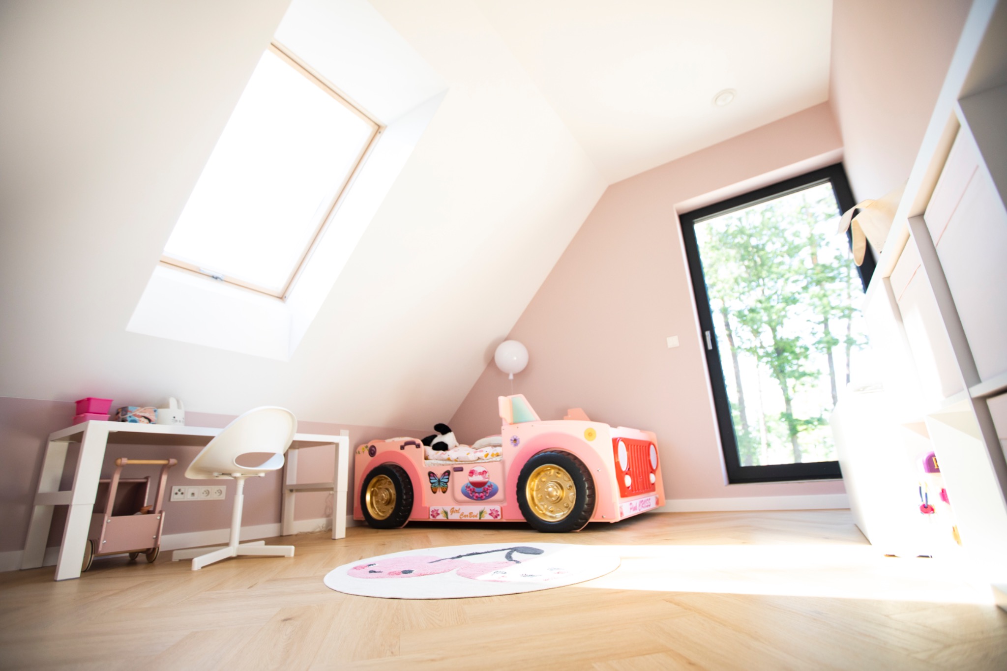 Pokój dziecięcy na poddaszu z łóżkiem w kształcie różowego samochodu, biurkiem i oknem dachowym. Jasne drewniane podłogi i pastelowe ściany tworzą przytulną atmosferę.