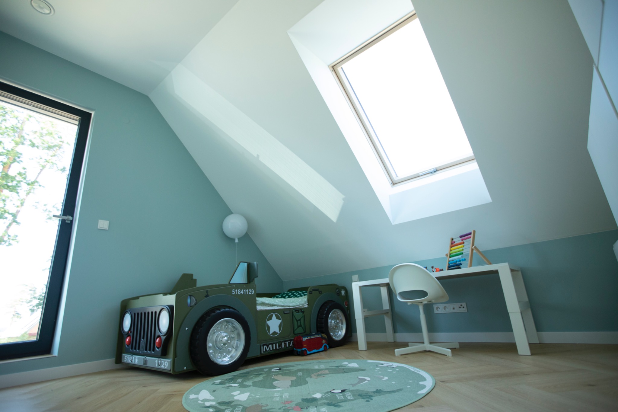 Pokój dziecięcy na poddaszu z oknem dachowym, pomalowany na pastelowy błękit. Uwagę przykuwa łóżko w kształcie wojskowego jeepa i okrągły dywan z mapą świata.