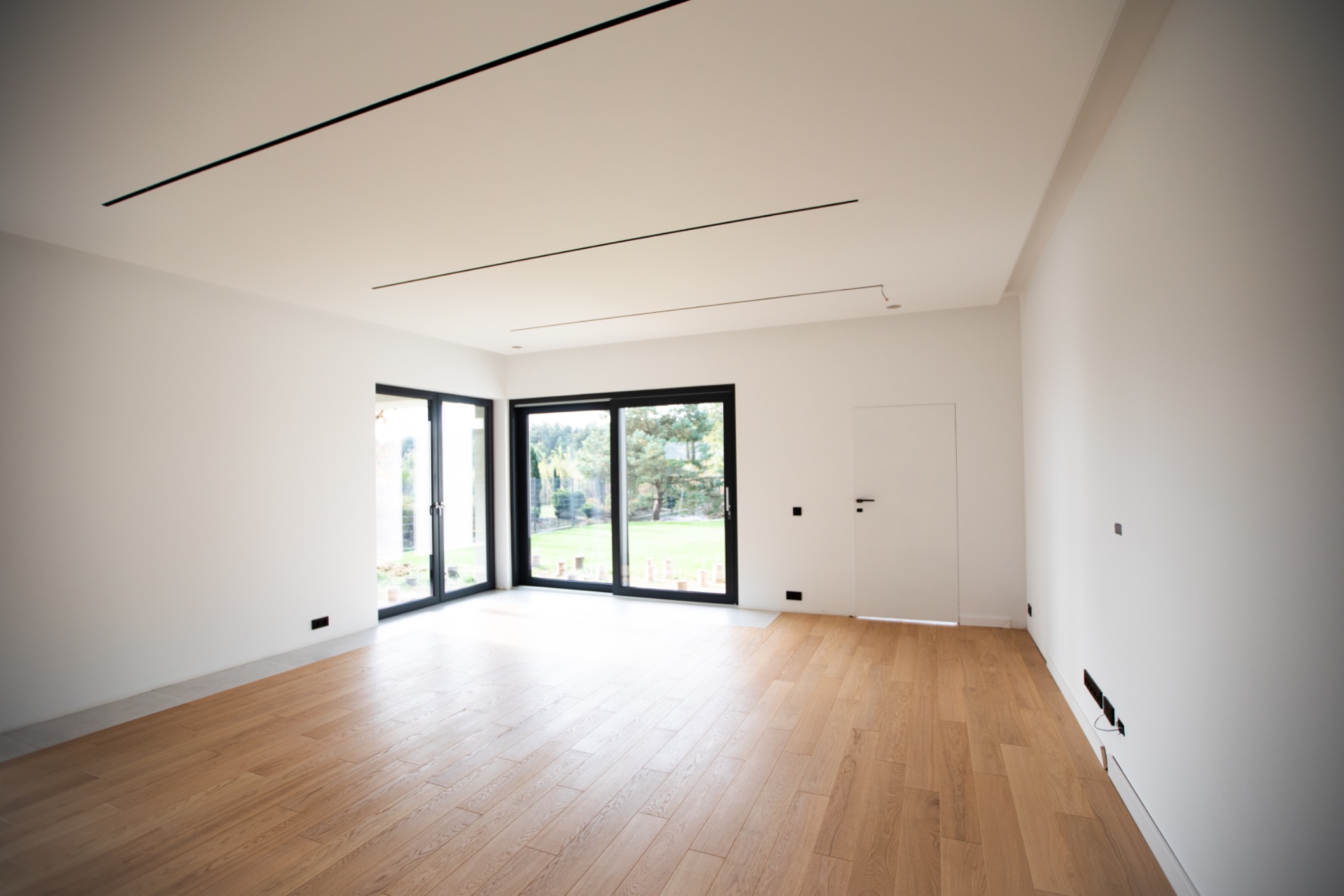 Jasne, przestronne wnętrze z drewnianą podłogą i dużymi oknami balkonowymi z czarnymi ramami. Minimalistyczne oświetlenie sufitowe. Świeżo wykończone ściany.