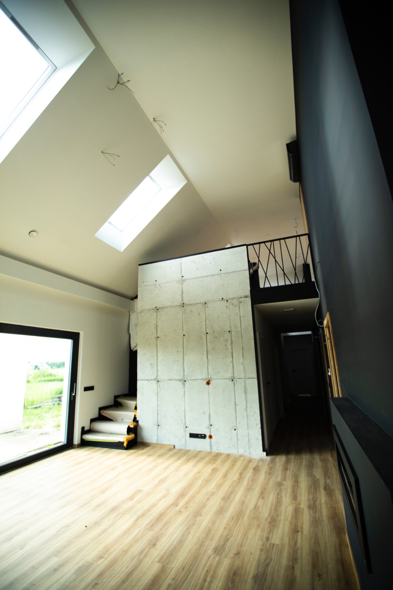 Jasne wnętrze z drewnianą podłogą i betonową ścianą. Schody zabezpieczone folią. Okno dachowe wpuszcza światło. Minimalistyczny design z czarnymi akcentami.