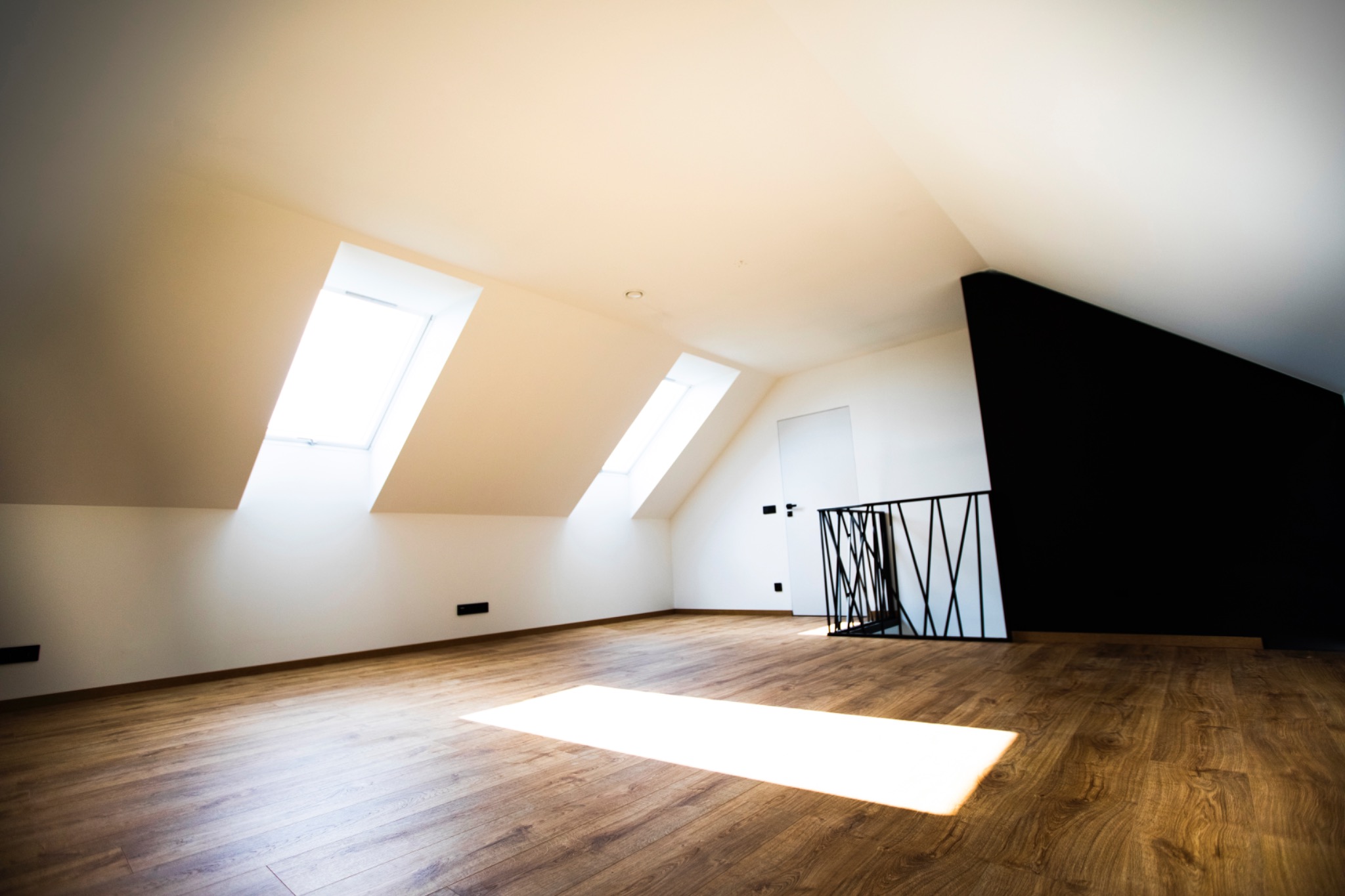 Jasne poddasze z drewnianą podłogą, oświetlone przez okna dachowe. Czarna ściana i nowoczesna balustrada dodają kontrastu. Widoczny fragment drzwi.
