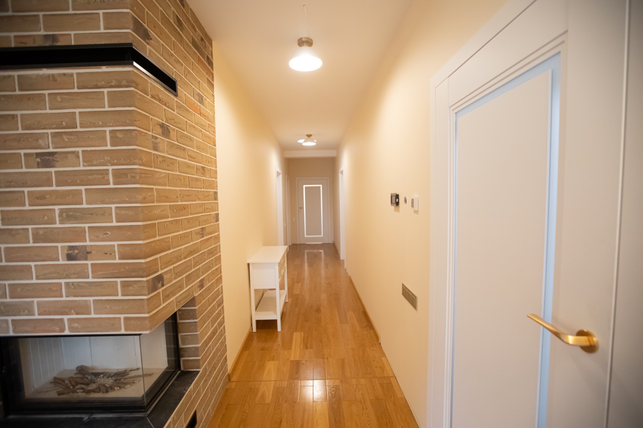 Wykończony korytarz z ceglanym kominkiem, drewnianą podłogą i białymi drzwiami. Widok w głąb korytarza z oświetleniem sufitowym i białymi ścianami. Nowoczesny design.