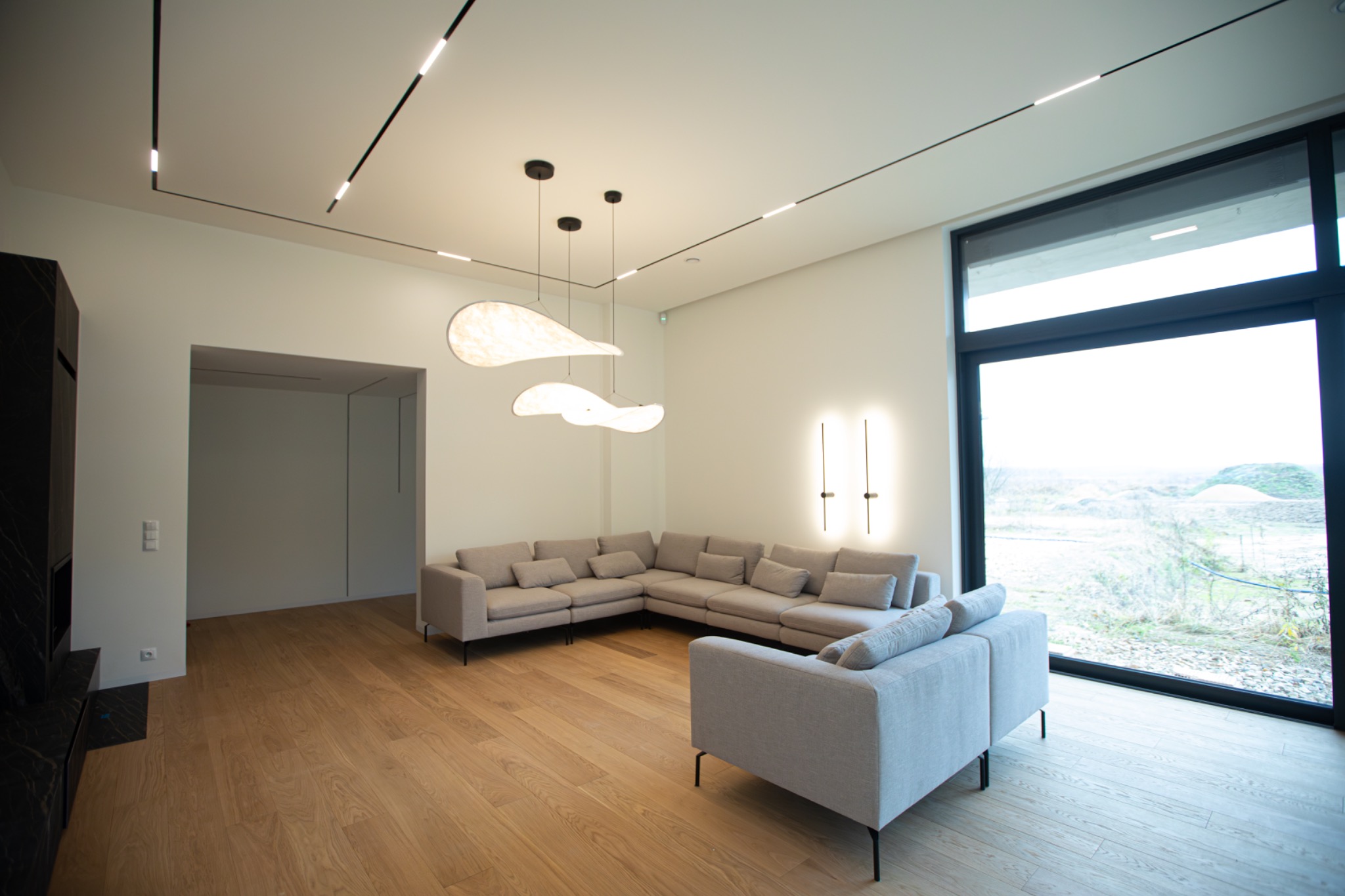 Jasne wnętrze salonu z parkietem, narożną sofą i dużym oknem z widokiem na zewnątrz. Minimalistyczne oświetlenie sufitowe i lampy wiszące dodają nowoczesnego charakteru.