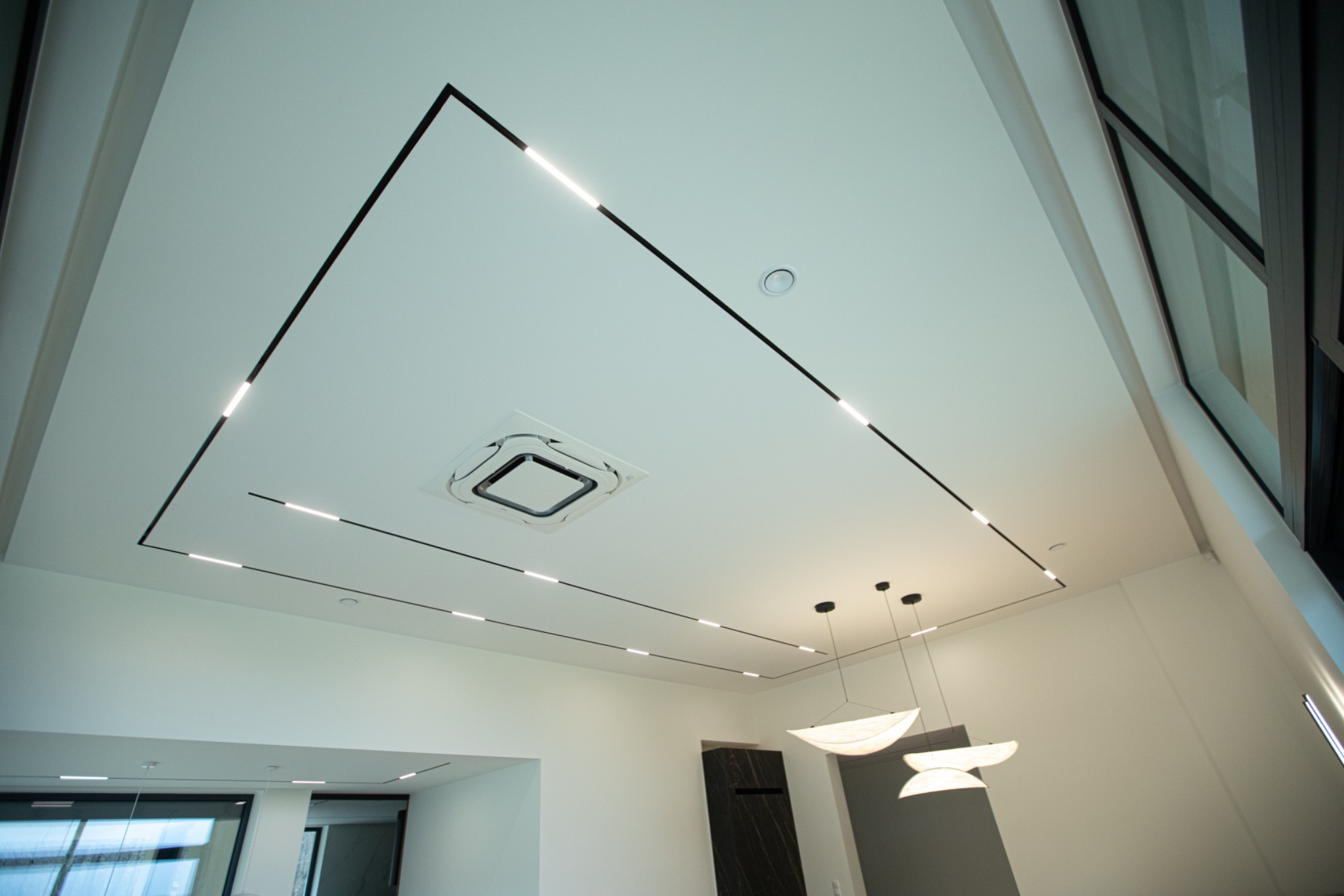 Nowoczesny sufit podwieszany z liniowym oświetleniem LED i klimatyzacją, minimalistyczne lampy wiszące, jasne wnętrze z dużymi oknami.