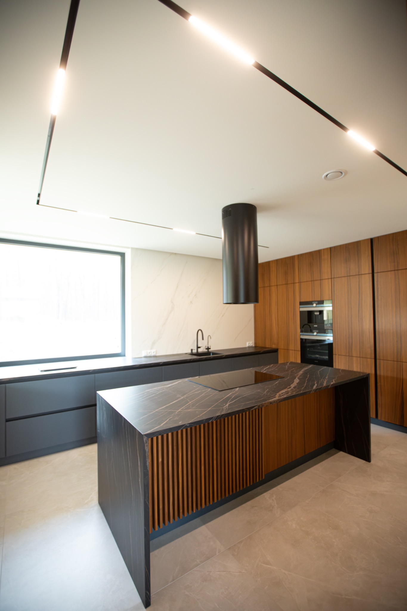 Nowoczesna kuchnia z wyspą o marmurowym blacie i drewnianymi akcentami. Minimalistyczne oświetlenie sufitowe i zabudowa kuchenna w odcieniach szarości i drewna.