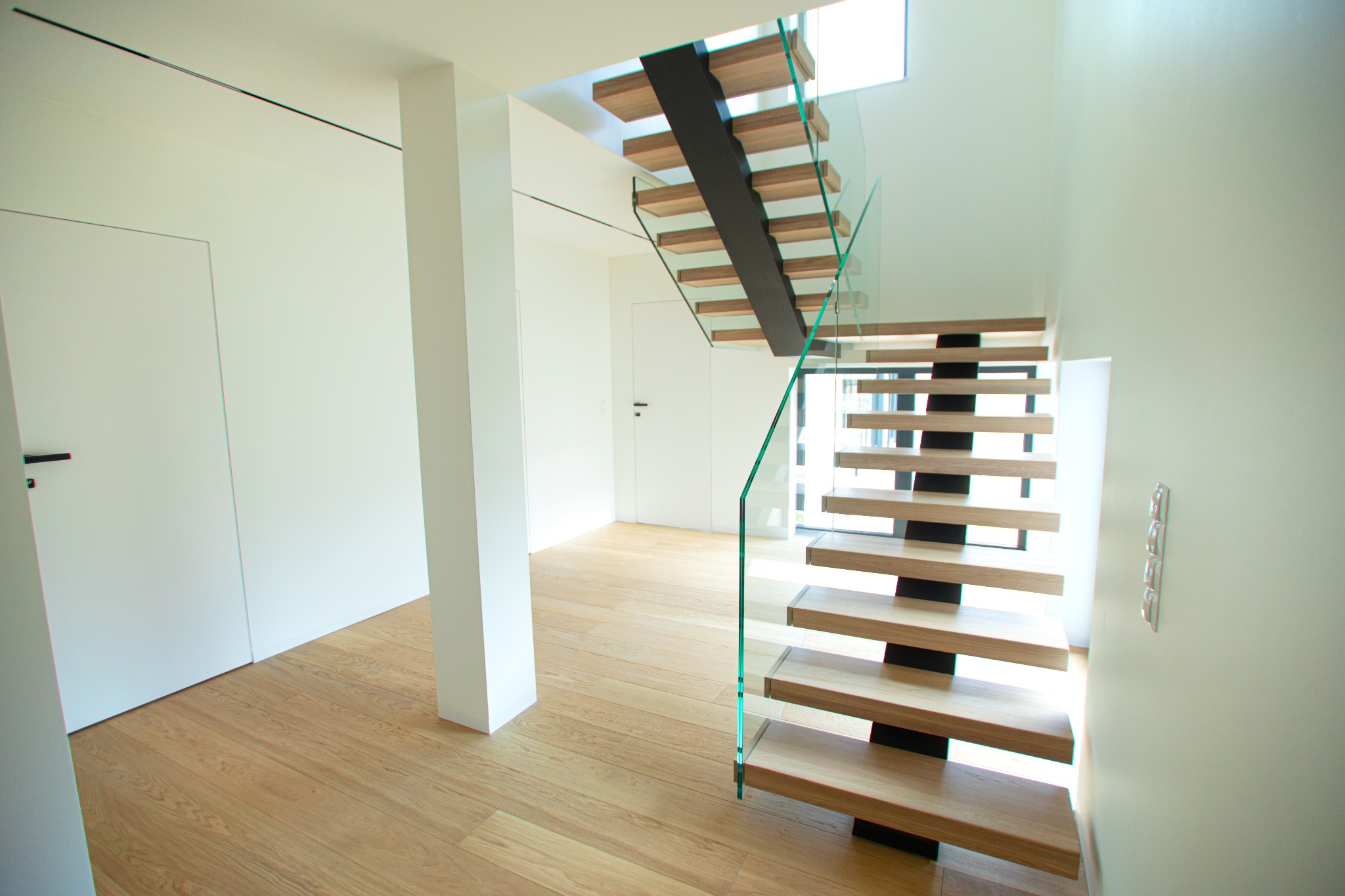 Nowoczesne, minimalistyczne schody z drewnianymi stopniami i szklaną balustradą w jasnym wnętrzu. Widoczna drewniana podłoga i białe ściany z drzwiami.