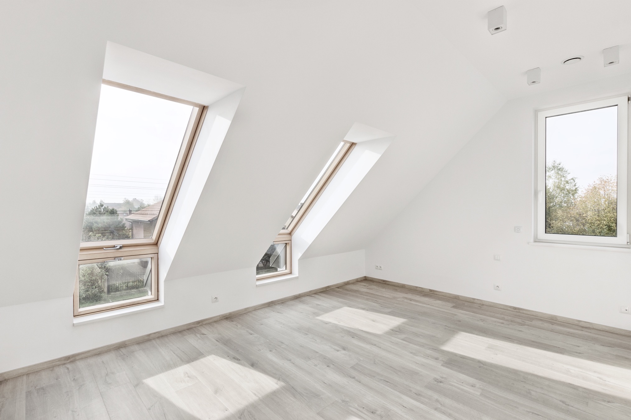 Jasne poddasze z drewnianą podłogą i oknami dachowymi. Dwa okna dachowe pod skosem i jedno pionowe. Białe ściany i sufit. Minimalistyczny design.