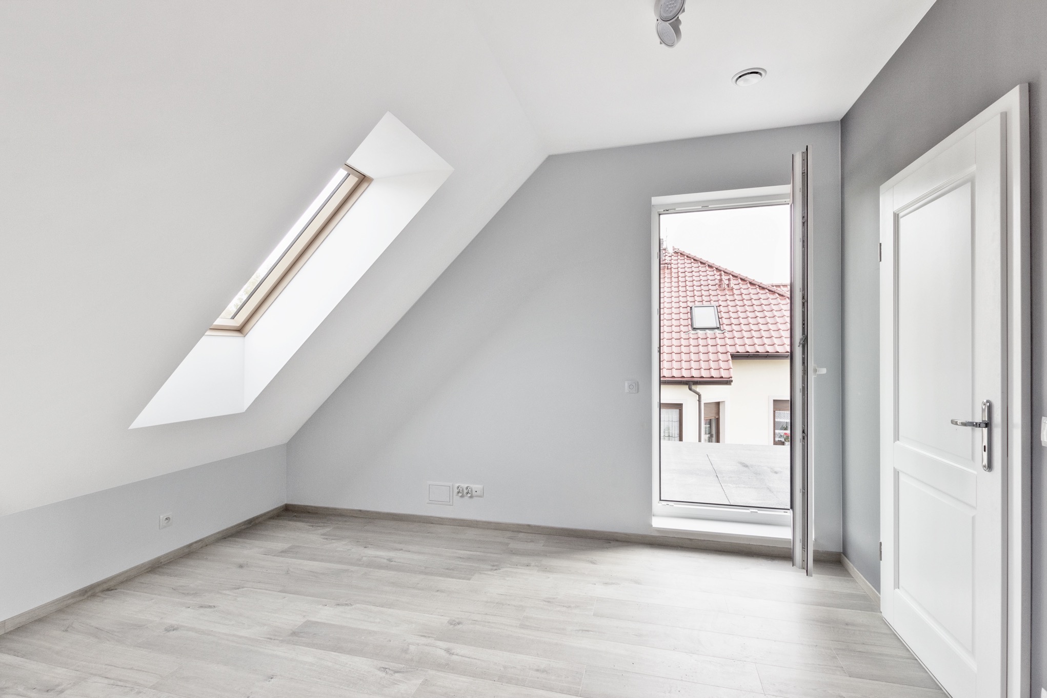 Jasne poddasze z oknem dachowym i otwartymi drzwiami balkonowymi. Ściany pomalowane na biało i szaro, podłoga wyłożona panelami w odcieniach szarości.