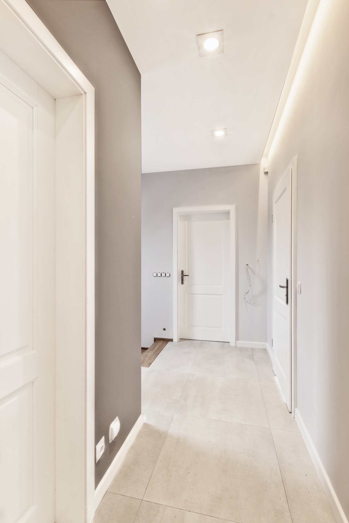 Jasny korytarz po remoncie z białymi drzwiami, szarą ścianą akcentującą i kremową podłogą z płytek. Oświetlenie sufitowe punktowe i listwowe.
