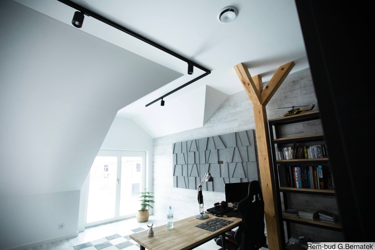 Jasne poddasze ze skośnym sufitem i oknem. Ściana z betonową dekoracją, biurko z lampką i komputerem. Czarny system oświetlenia szynowego na suficie.