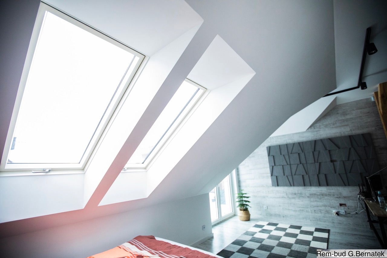 Jasne poddasze z oknami dachowymi i dekoracyjną ścianą. Uwagę przykuwa geometryczny wzór na ścianie i szachownica na dywanie. Widoczny fragment łóżka i roślina doniczkowa.