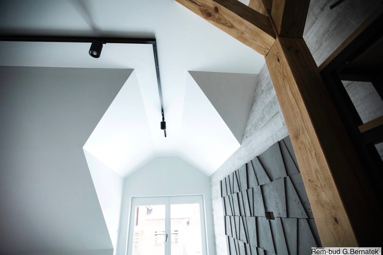 Nowoczesne wykończenie wnętrza: białe ściany, drewniana belka stropowa i dekoracyjna ściana z geometrycznych paneli w odcieniach szarości. Oświetlenie punktowe.