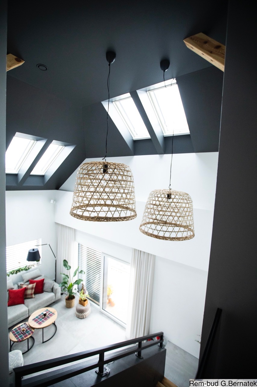 Nowoczesne wnętrze z antresolą: szara ściana, okna dachowe i wiszące lampy z naturalnego materiału. Widok z góry na salon z sofą i stolikami.