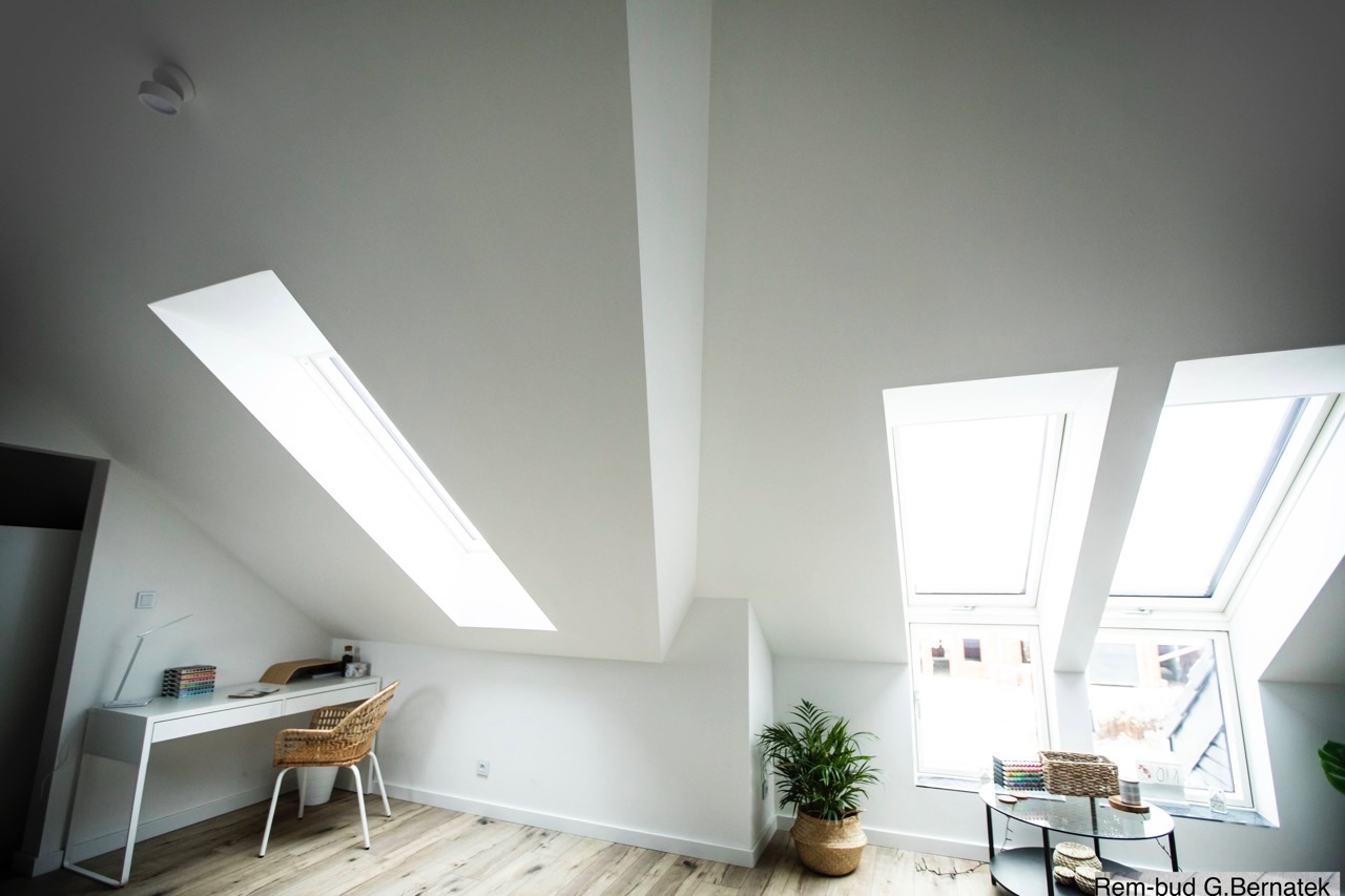 Jasne poddasze ze skośnymi ścianami i oknami dachowymi, biurko z wiklinowym krzesłem, roślina doniczkowa. Minimalistyczny design, drewniana podłoga.