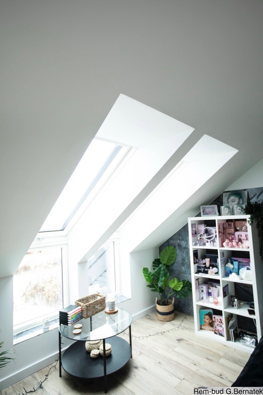 Jasne poddasze z oknami dachowymi, wykończone w bieli. Na podłodze panele, stolik kawowy i regał z dekoracjami. Widok na nowoczesne wnętrze po remoncie.