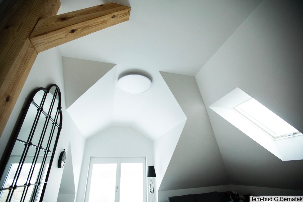 Białe wnętrze poddasza z drewnianą belką, oknem dachowym i lustrem w stylu gotyckim. Widoczne oświetlenie sufitowe i fragment sofy. Ujęcie z dołu.