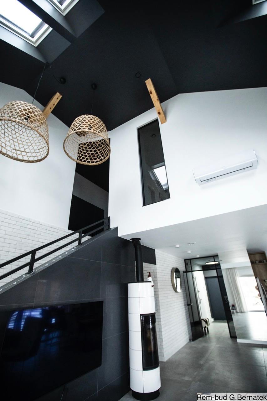 Nowoczesne wnętrze z czarno-białymi akcentami: schody z ciemnej glazury, biała cegła, wiszące lampy z wikliny i czarny sufit. Minimalistyczny design z kominkiem.