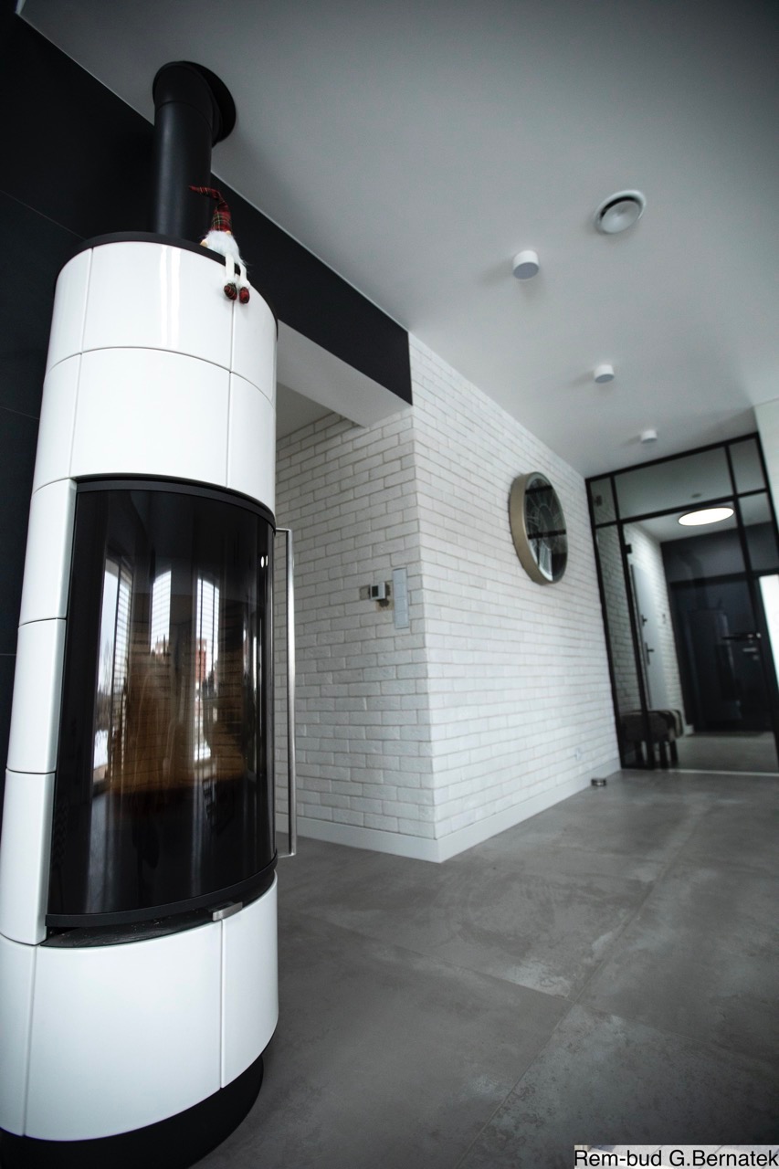 Nowoczesne wykończenie wnętrza: biała cegła na ścianie, szary beton na podłodze i designerski piec kaflowy z czarnym kominem. Minimalistyczny styl.