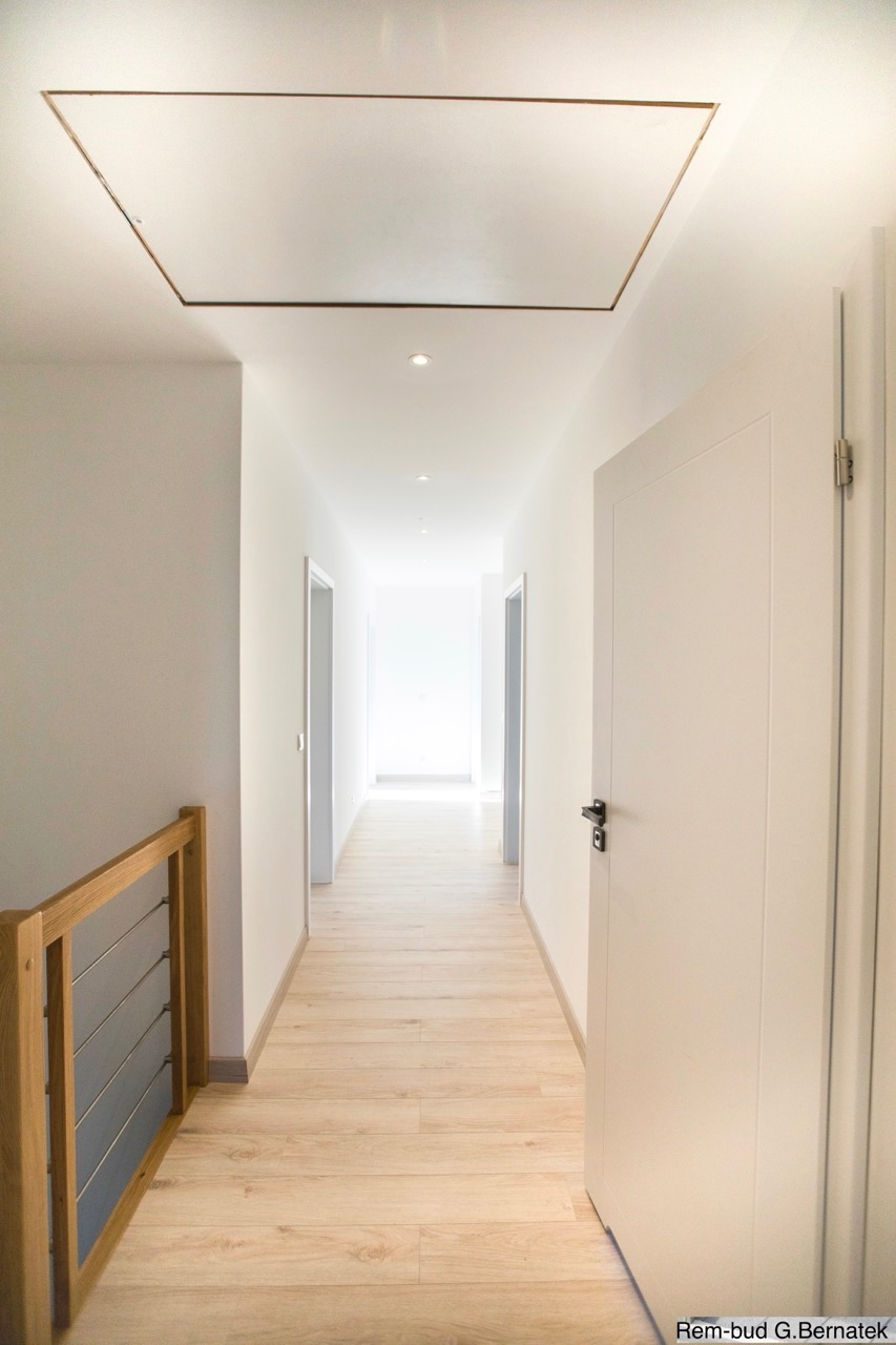 Jasny korytarz po remoncie: panele podłogowe, białe ściany i drzwi. Widoczna klapa rewizyjna w suficie i fragment balustrady. Perspektywa długa, z oświetleniem punktowym.