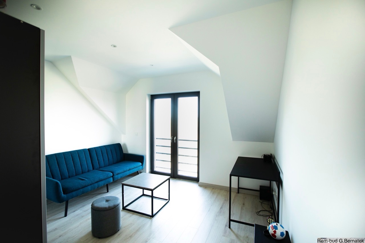 Jasne poddasze po remoncie: błękitna sofa, stolik kawowy, biurko, okno balkonowe. Ściany i sufit białe, podłoga drewniana. Minimalistyczny styl wykończenia wnętrza.