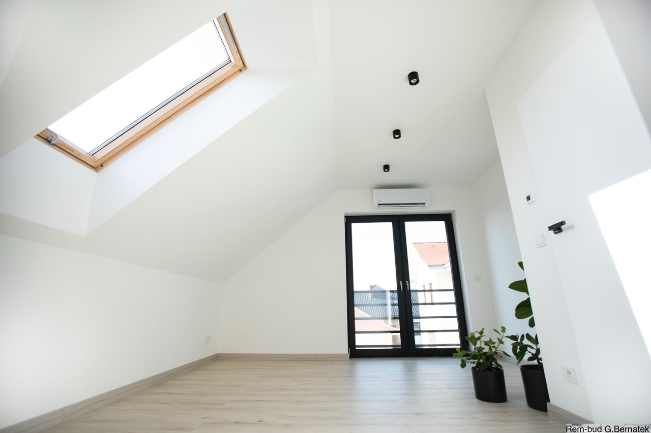 Jasne poddasze po remoncie: białe ściany, okno dachowe, parkiet, czarne lampy i drzwi balkonowe. Minimalistyczny styl, roślina doniczkowa w rogu.