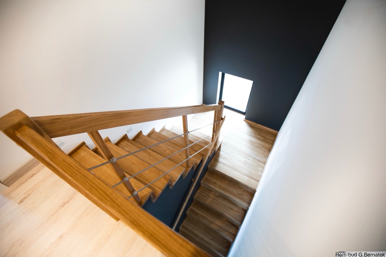 Drewniane schody z metalową balustradą, widok z góry. Białe i grafitowe ściany, jasna podłoga. Nowoczesne wykończenie wnętrza z oknem w tle.