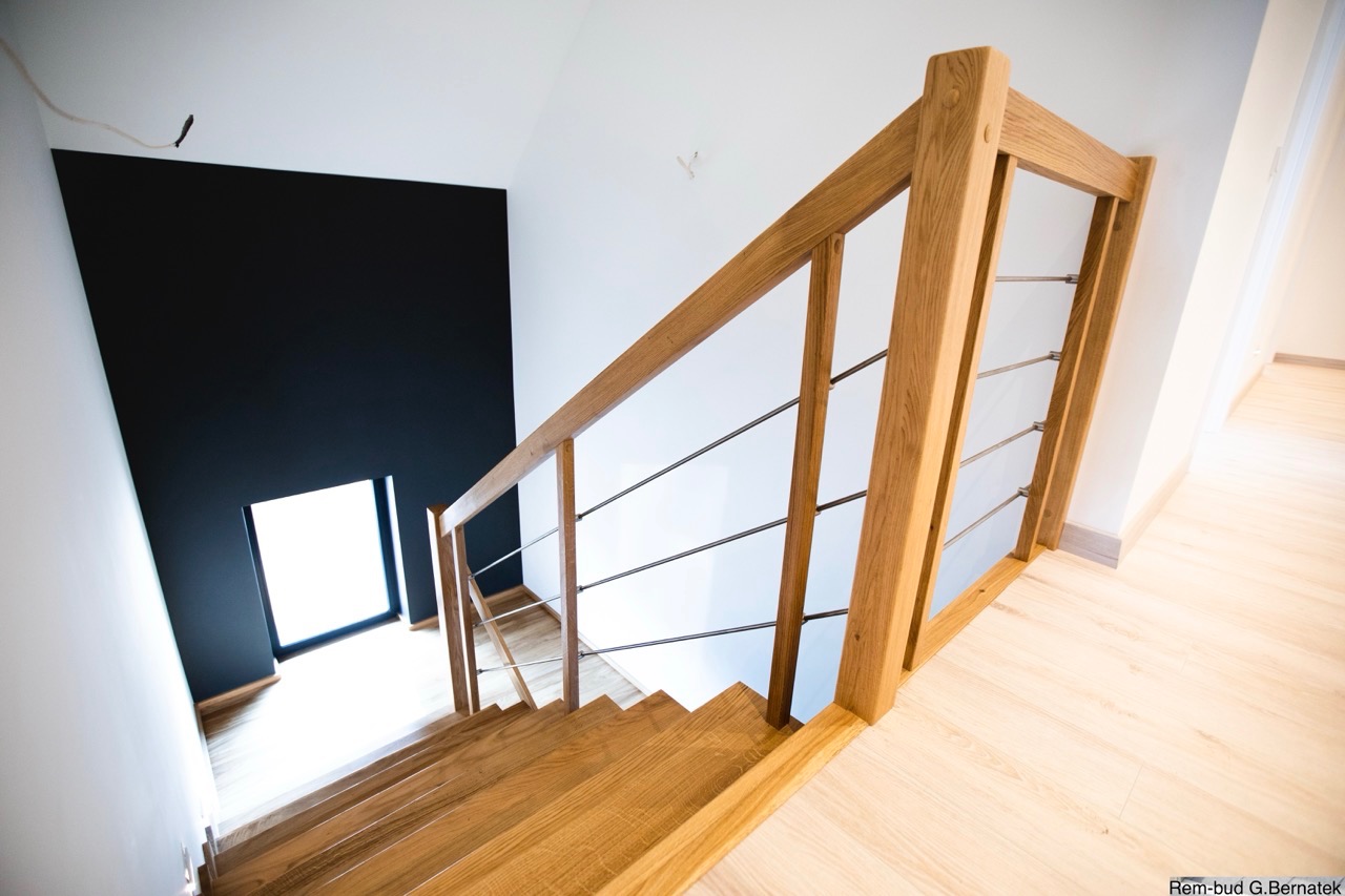 Drewniane schody z metalową balustradą w nowoczesnym wnętrzu. Kontrast bieli i czerni na ścianach. Widok z góry na jasną podłogę i okno w tle.