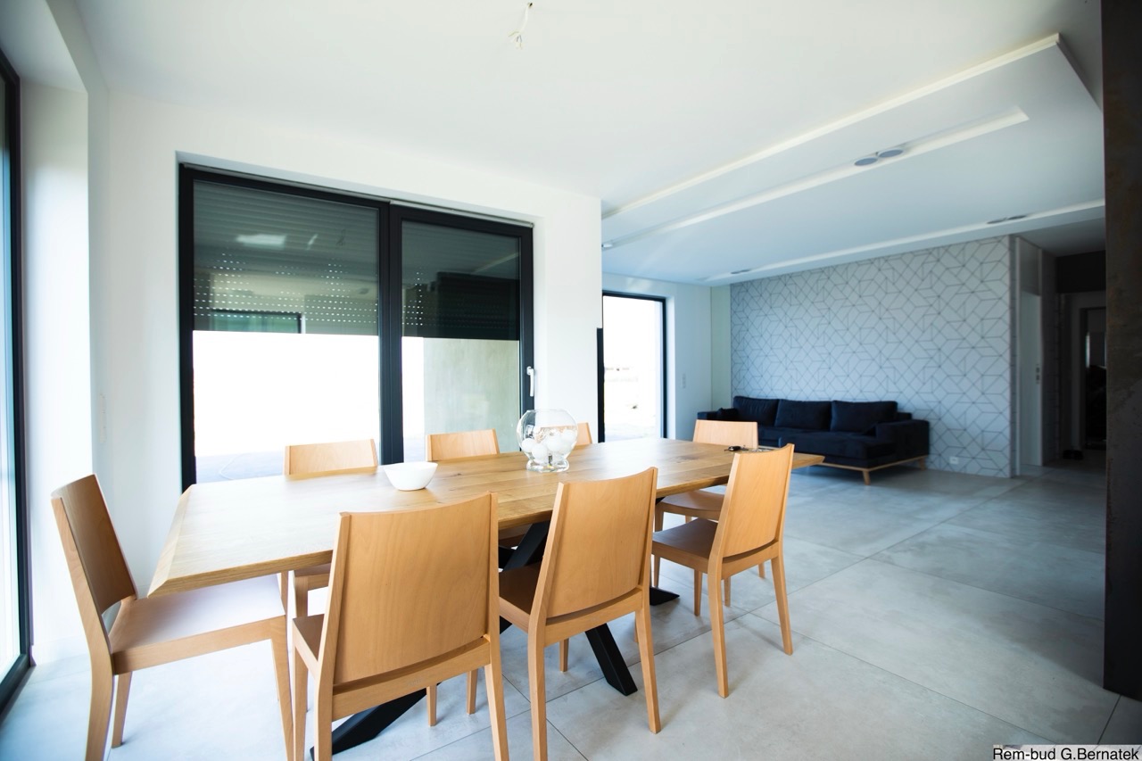 Jasne wnętrze jadalni w Gliwicach: drewniany stół z krzesłami, szara ściana z geometrycznym wzorem, czarna sofa. Nowoczesny design i minimalistyczny styl.