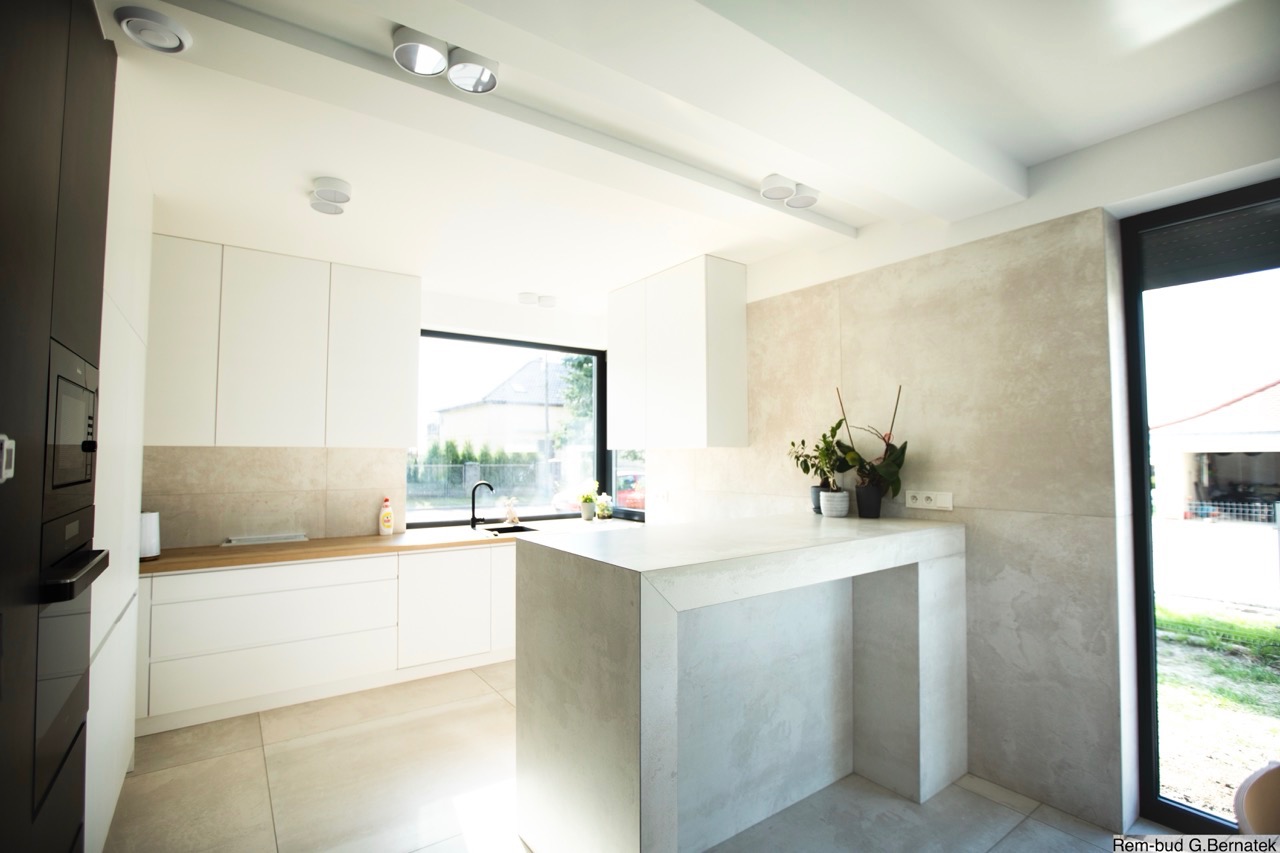 Jasna kuchnia z białymi szafkami i wyspą wykończoną w odcieniach szarości. Minimalistyczny design, duże okno z widokiem na zieleń. Nowoczesne oświetlenie sufitowe.