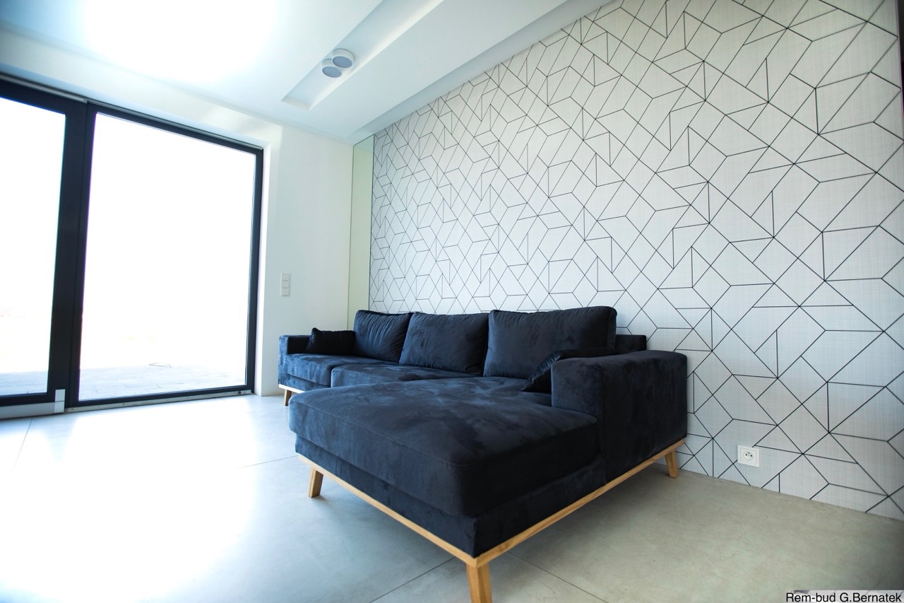 Nowoczesny salon z geometryczną tapetą i narożną sofą w odcieniach granatu. Duże okno z czarną ramą wpuszcza dużo światła. Minimalistyczne wykończenie.