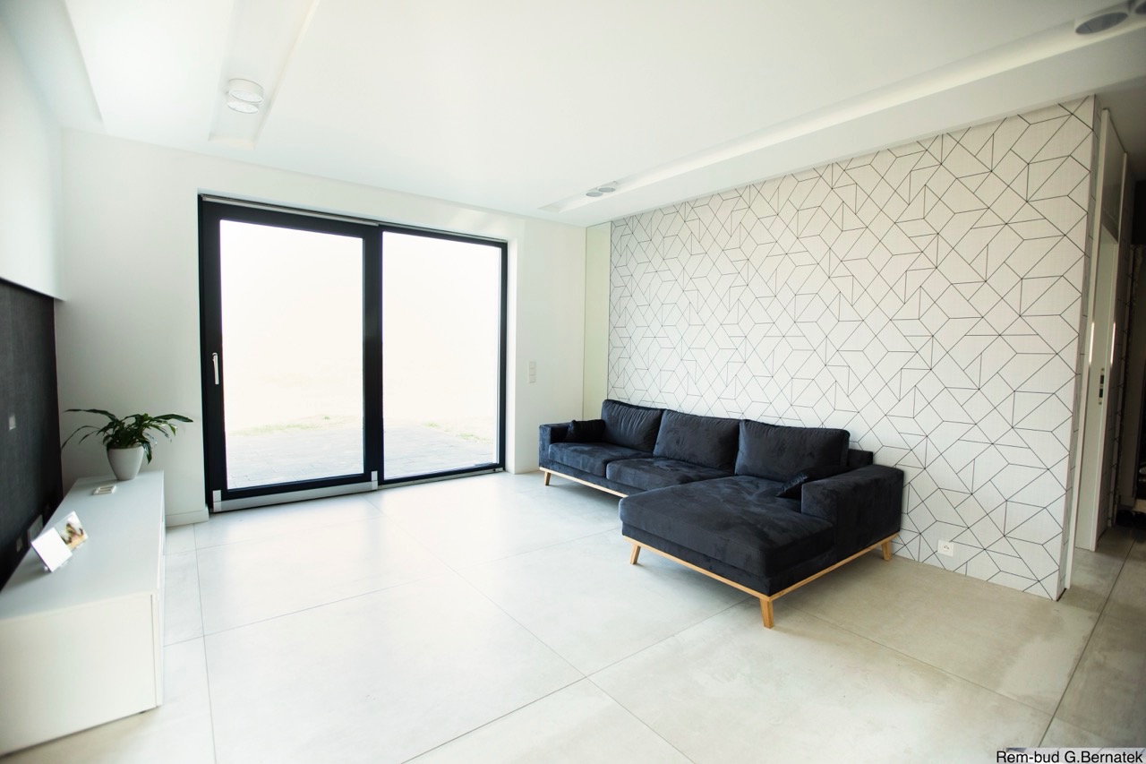 Minimalistyczne wnętrze: czarna sofa narożna na tle ściany z geometrycznym wzorem, duże okno przesuwne, białe płytki podłogowe i szafka RTV z rośliną.