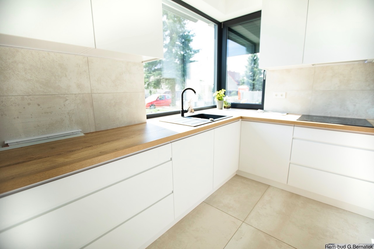 Nowoczesna, jasna kuchnia z białymi szafkami, drewnianym blatem i czarną armaturą. Duże okno z widokiem na zieleń. Beżowe płytki na podłodze i ścianie.