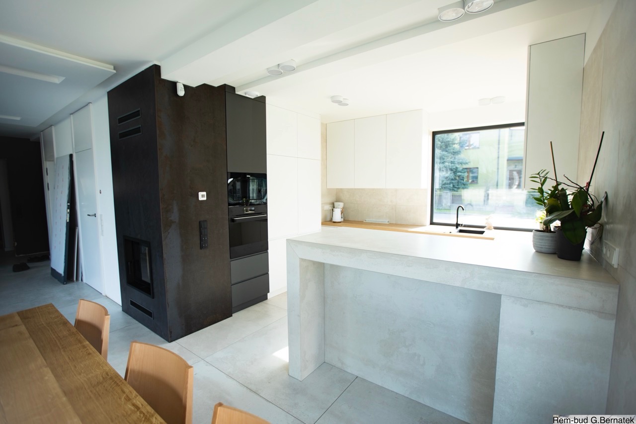 Nowoczesna kuchnia z wyspą w odcieniach bieli, szarości i czerni. Minimalistyczne szafki, duże okno z widokiem na zieleń. Elementy wykończenia imitujące beton.