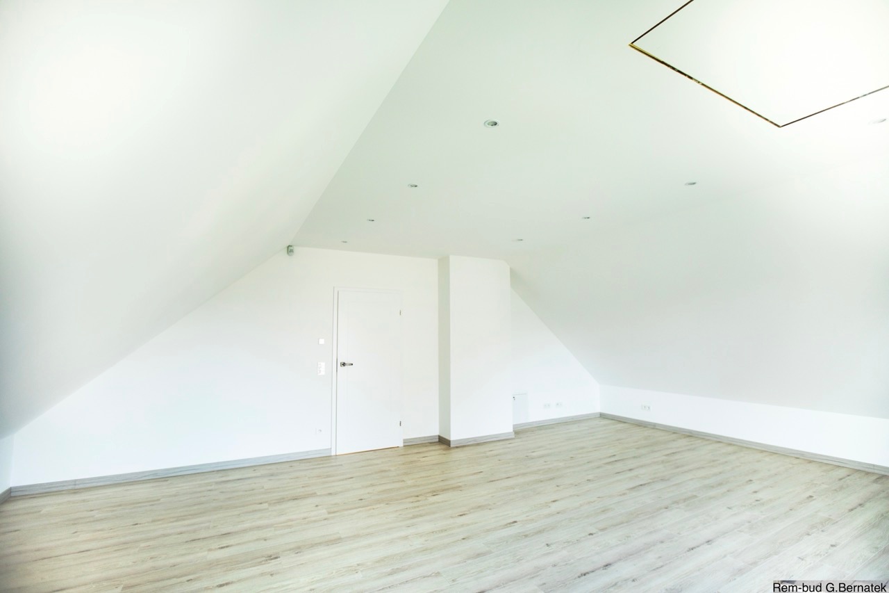 Jasne, świeżo wykończone poddasze z białymi ścianami i panelami podłogowymi w odcieniach szarości. Widoczne skosy, białe drzwi i oświetlenie punktowe.