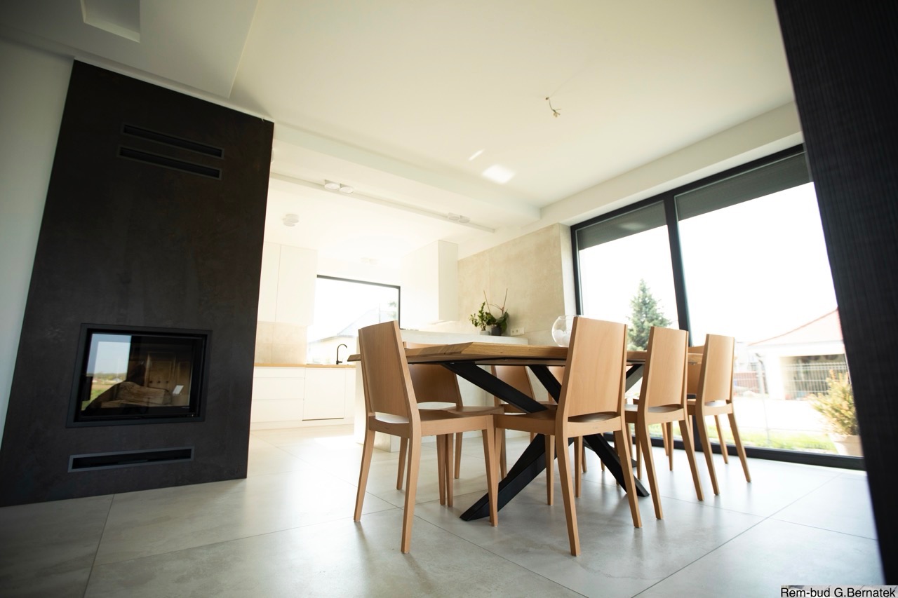 Jasne wnętrze jadalni z drewnianym stołem i krzesłami, nowoczesny kominek z czarną obudową, duże okno z widokiem na zewnątrz, szara podłoga z płytek.