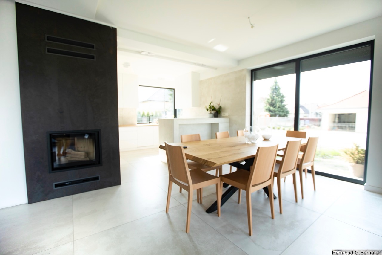 Jasne wnętrze z kominkiem o ciemnej obudowie i drewnianym stołem. Minimalistyczny design, duże okno z widokiem na zieleń. Nowoczesne wykończenie.