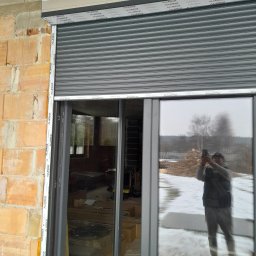 Szara roleta zewnętrzna Aluplast zamontowana na oknie z widocznym odbiciem osoby robiącej zdjęcie. Surowy mur z cegły i betonowy strop w tle.