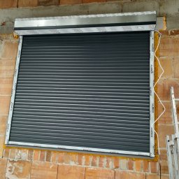 Szara roleta zewnętrzna zamontowana w oknie budynku z cegły, widoczne elementy montażowe i pianka uszczelniająca. Rama okienna z napisem 'ALUPROF'.