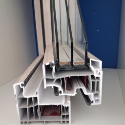 Justyna Pędziwiatr P&Home&Cars - Przekrój okna z trzema szybami, ukazujący wewnętrzną strukturę profilu z tworzywa sztucznego i uszczelki. Widoczne komory powietrzne i elementy mocujące.